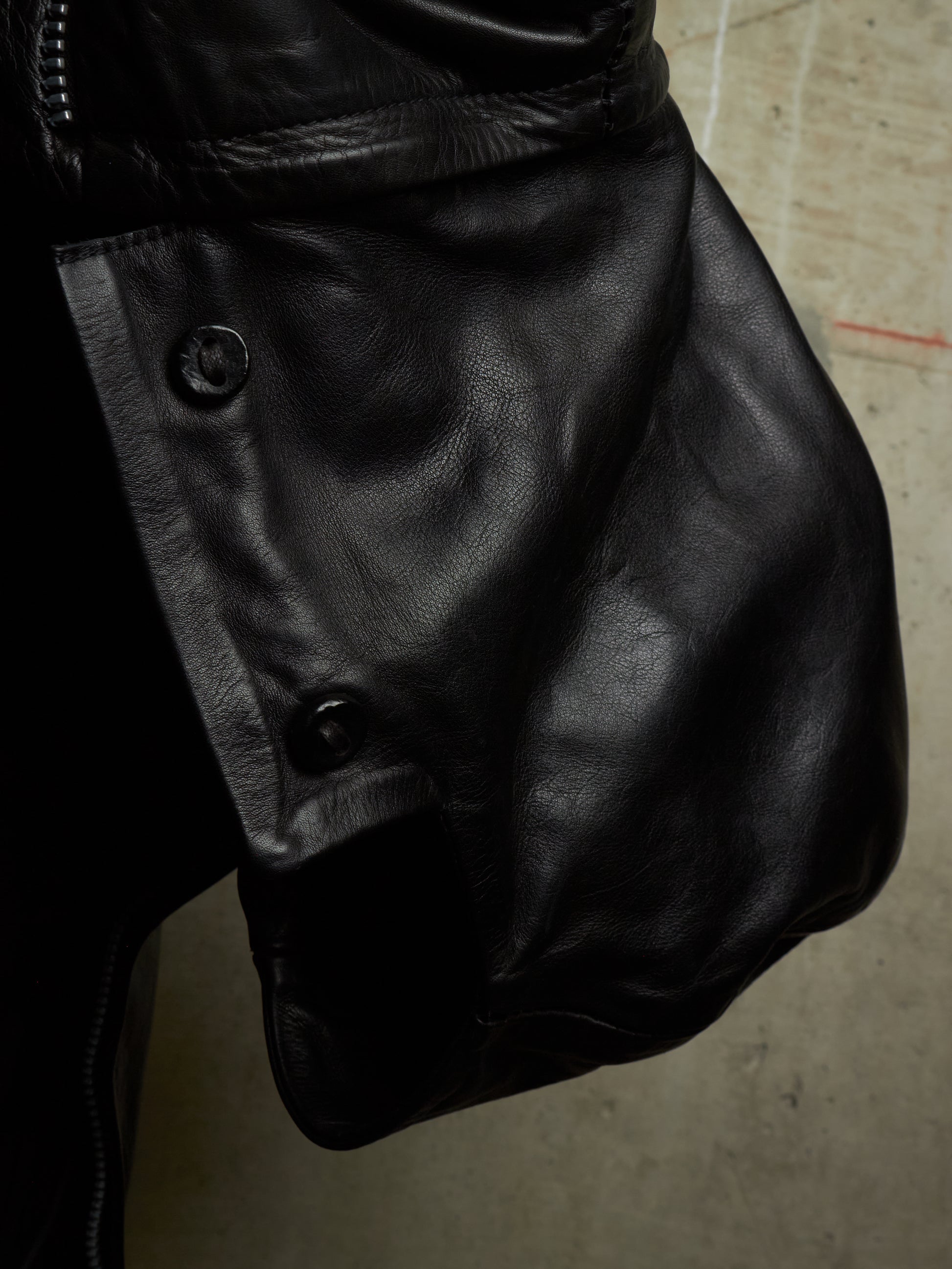 Close-up of a black leather jacket showing the side panel with snap buttons and a zipper, photographed against a concrete floor.