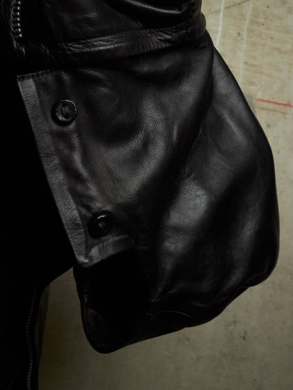 Close-up of a black leather jacket showing the side panel with snap buttons and a zipper, photographed against a concrete floor.