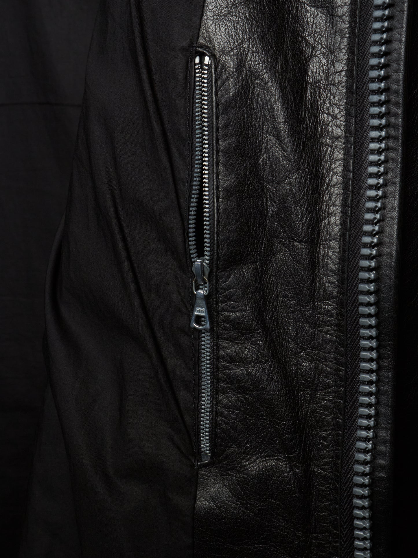 Close-up detail of a black jacket showing a vertical zippered pocket with matte black fabric on one side and textured black leather on the other, with a silver zipper pull.