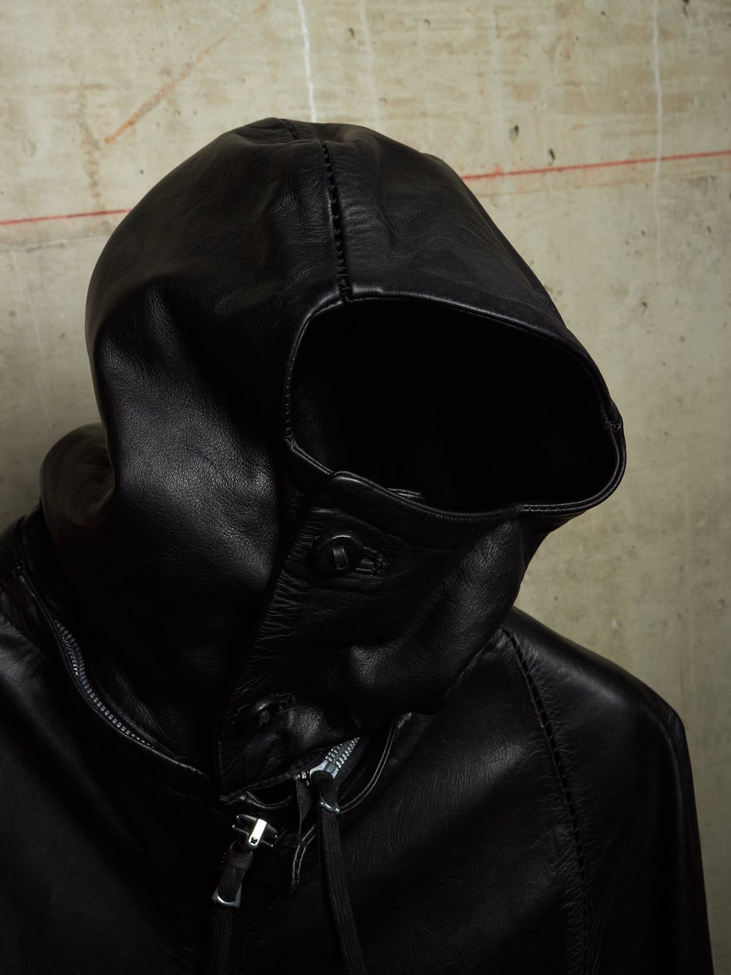 A close-up of a black leather hooded jacket with the hood pulled up, showing a dark button closure and zipper details, set against a textured concrete wall.