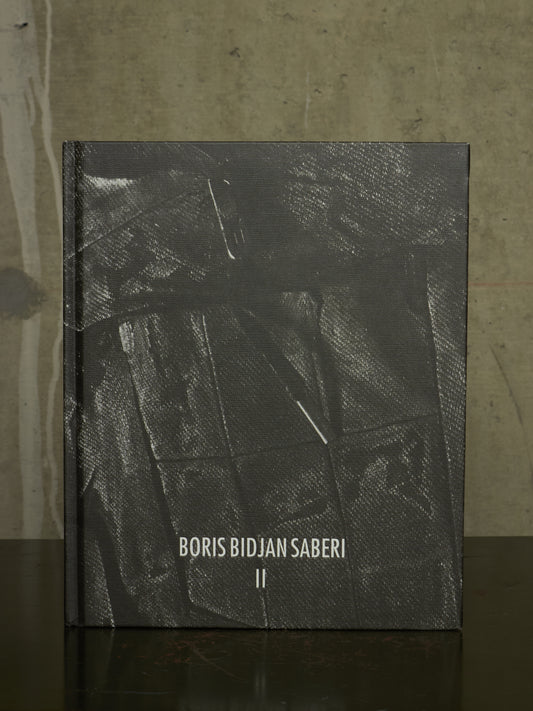 A hardcover book titled 'BORIS BIDJAN SABERI II' with a textured dark gray cover, standing upright on a dark reflective surface against a concrete-textured background.
