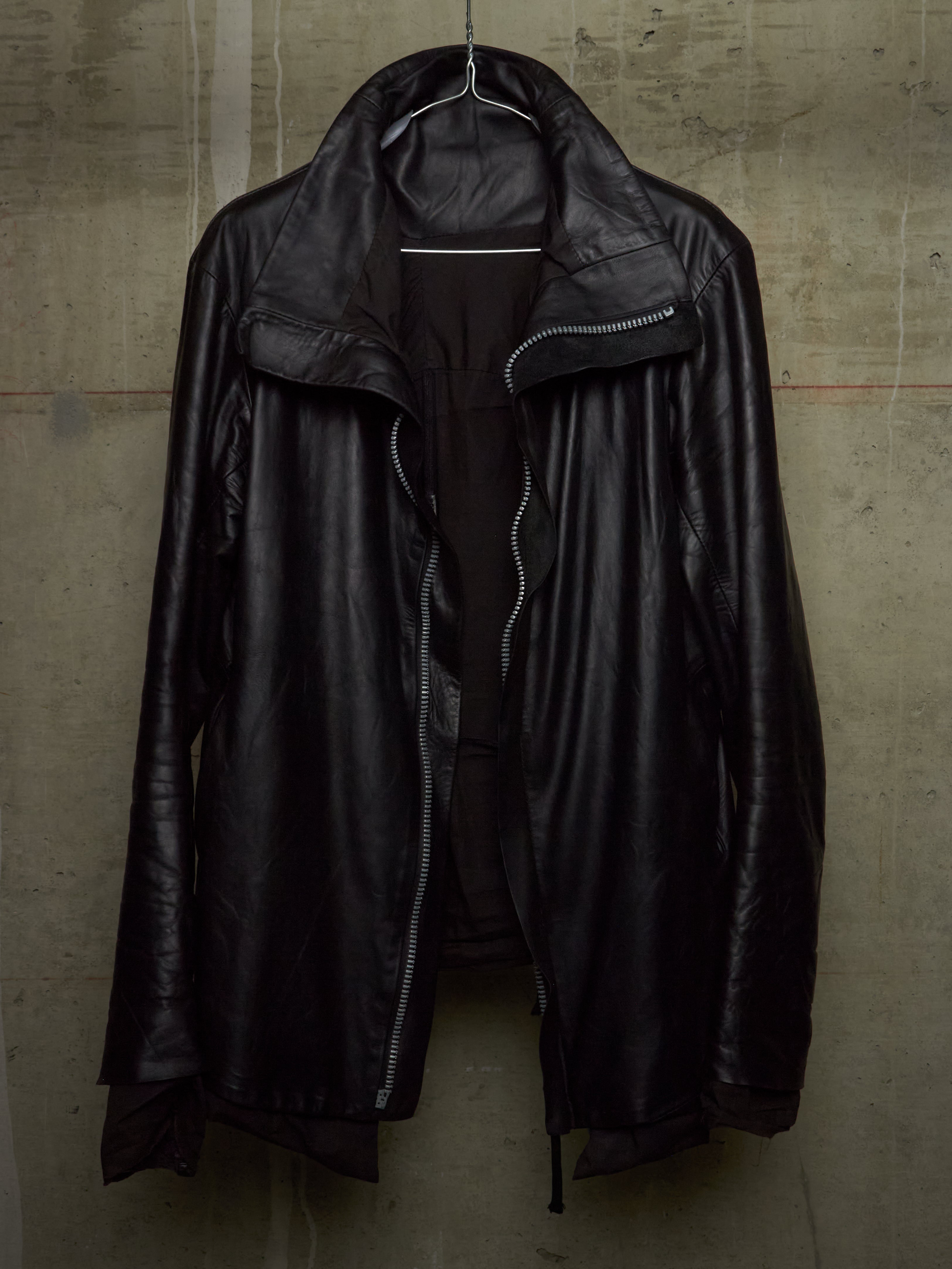 A black leather jacket with an asymmetrical zipper and high collar, hanging on a wire hanger against a textured concrete wall.