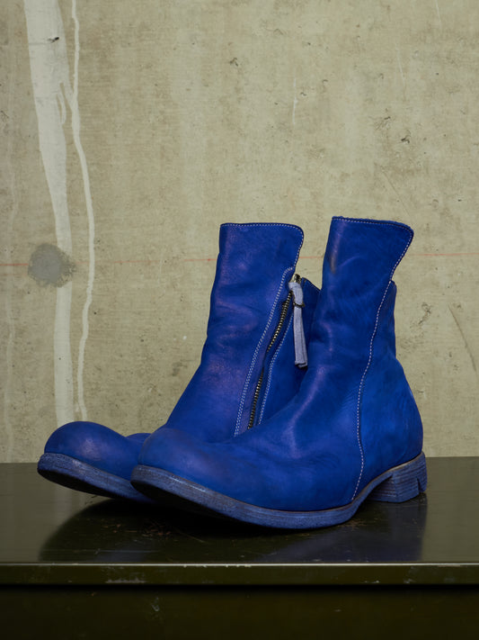 A pair of vibrant blue leather ankle boots with side zippers, displayed on a dark surface against a textured concrete wall.