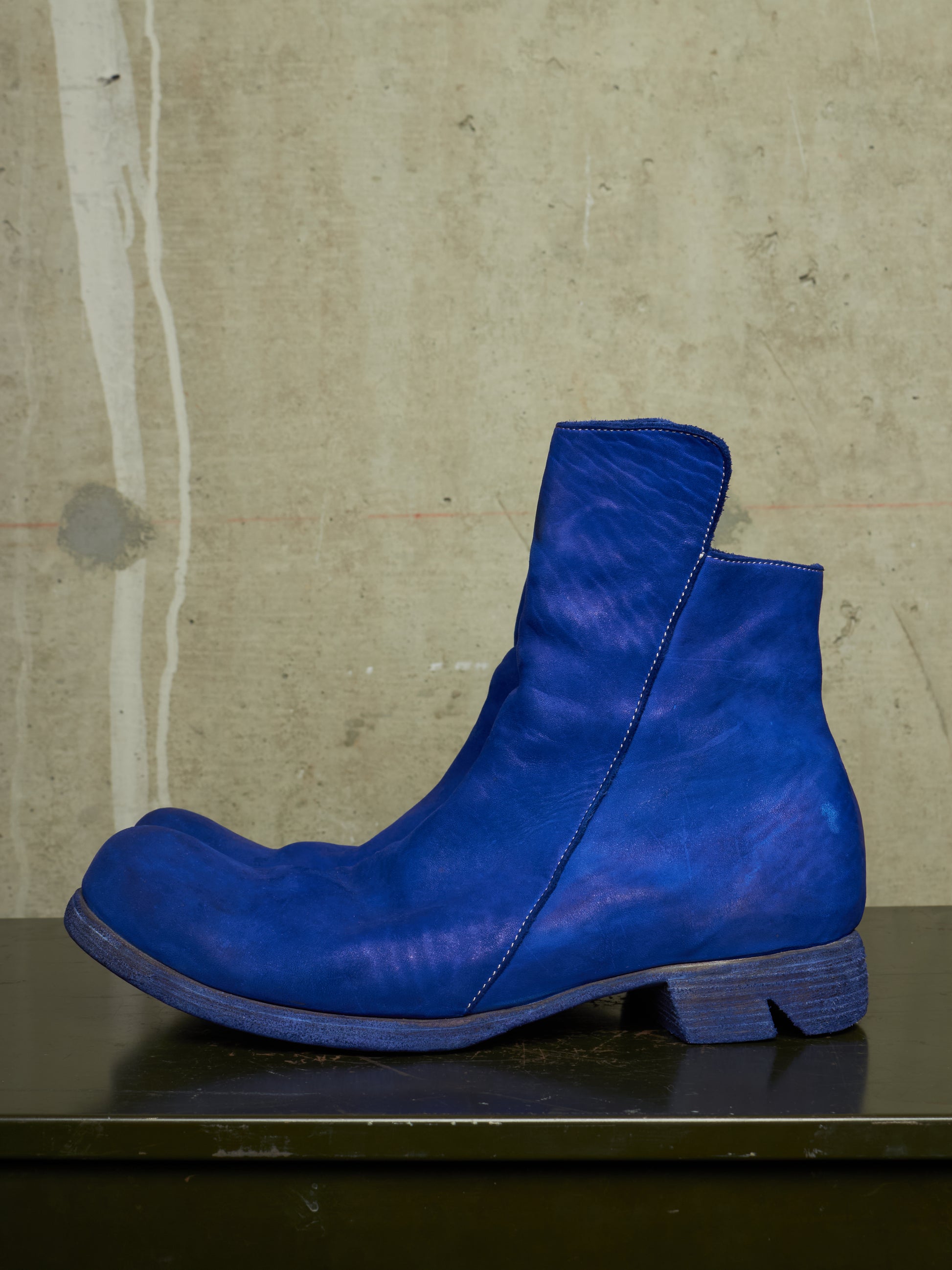 A single vibrant blue leather ankle boot with a chunky block heel, resting on a dark surface against a textured concrete wall.