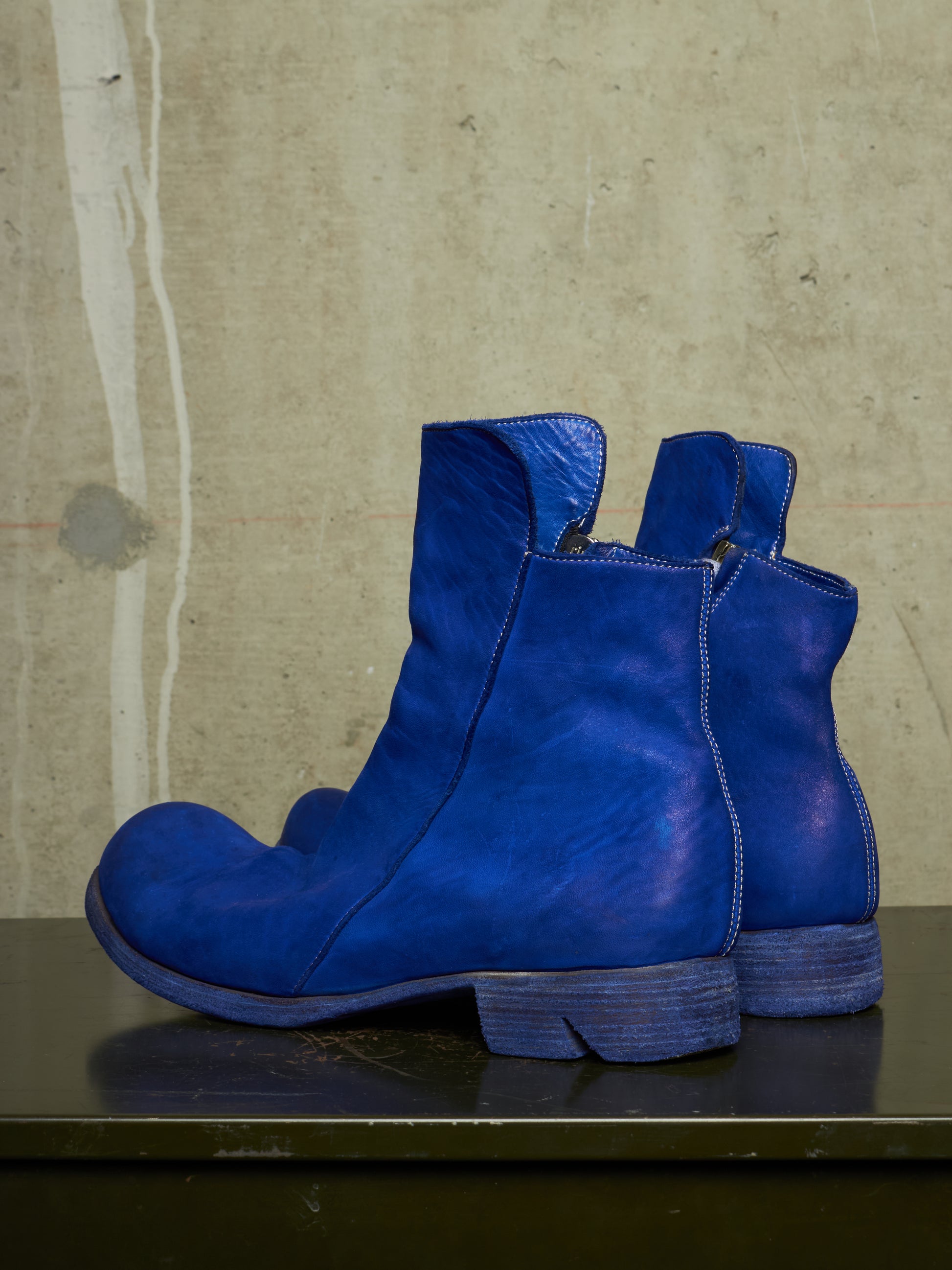 A pair of vibrant blue leather ankle boots with a distressed finish, displayed on a dark surface against a textured concrete wall.