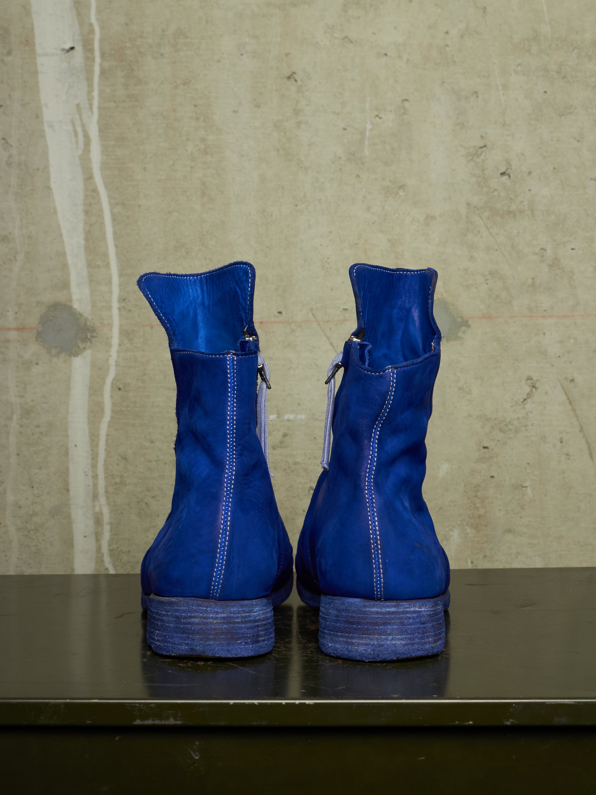 A pair of bright blue suede ankle boots with side zippers, shown from the back on a dark surface against a textured concrete wall.