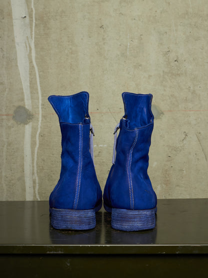 A pair of bright blue suede ankle boots with side zippers, shown from the back on a dark surface against a textured concrete wall.