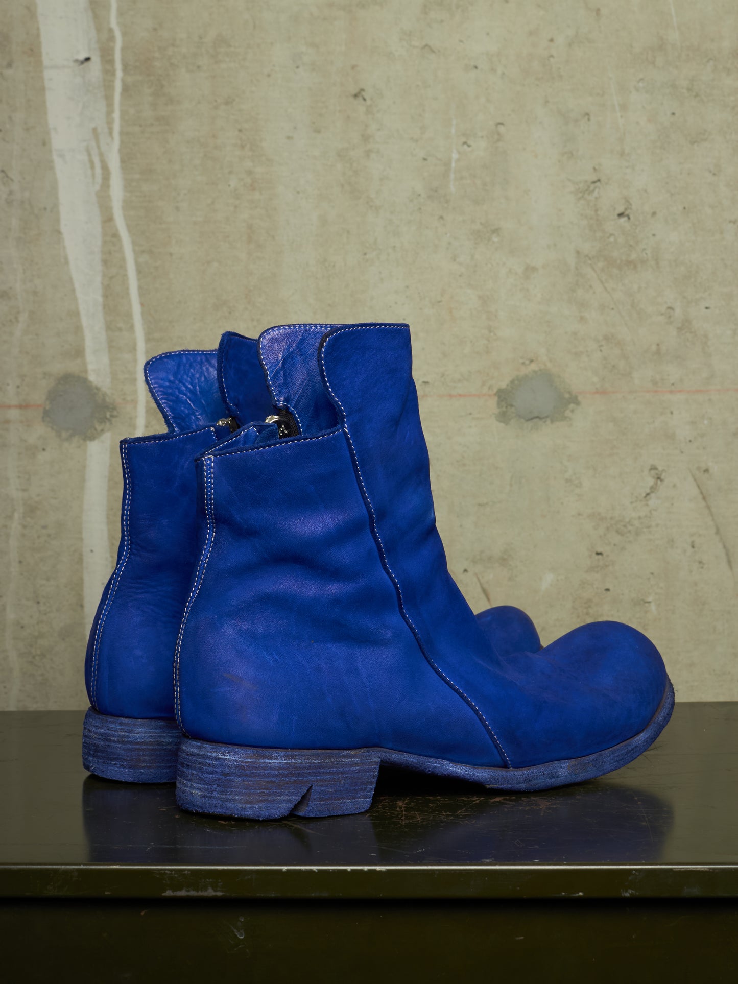 A pair of vibrant blue leather ankle boots with a low block heel, displayed on a dark surface against a textured concrete wall.