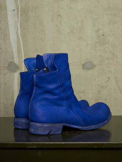 A pair of vibrant blue leather ankle boots with a low block heel, displayed on a dark surface against a textured concrete wall.