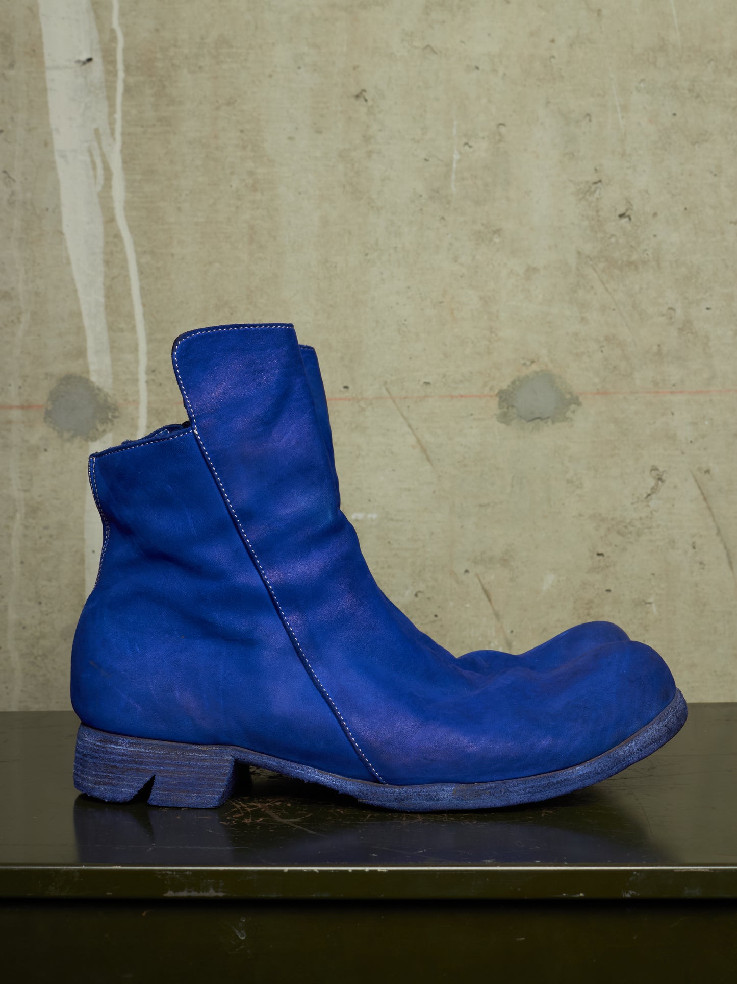 A single vibrant blue leather ankle boot with a low block heel, resting on a dark surface against a textured concrete wall.