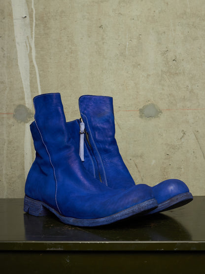 A pair of vibrant blue leather ankle boots with side zippers, displayed on a dark surface against a textured concrete wall.