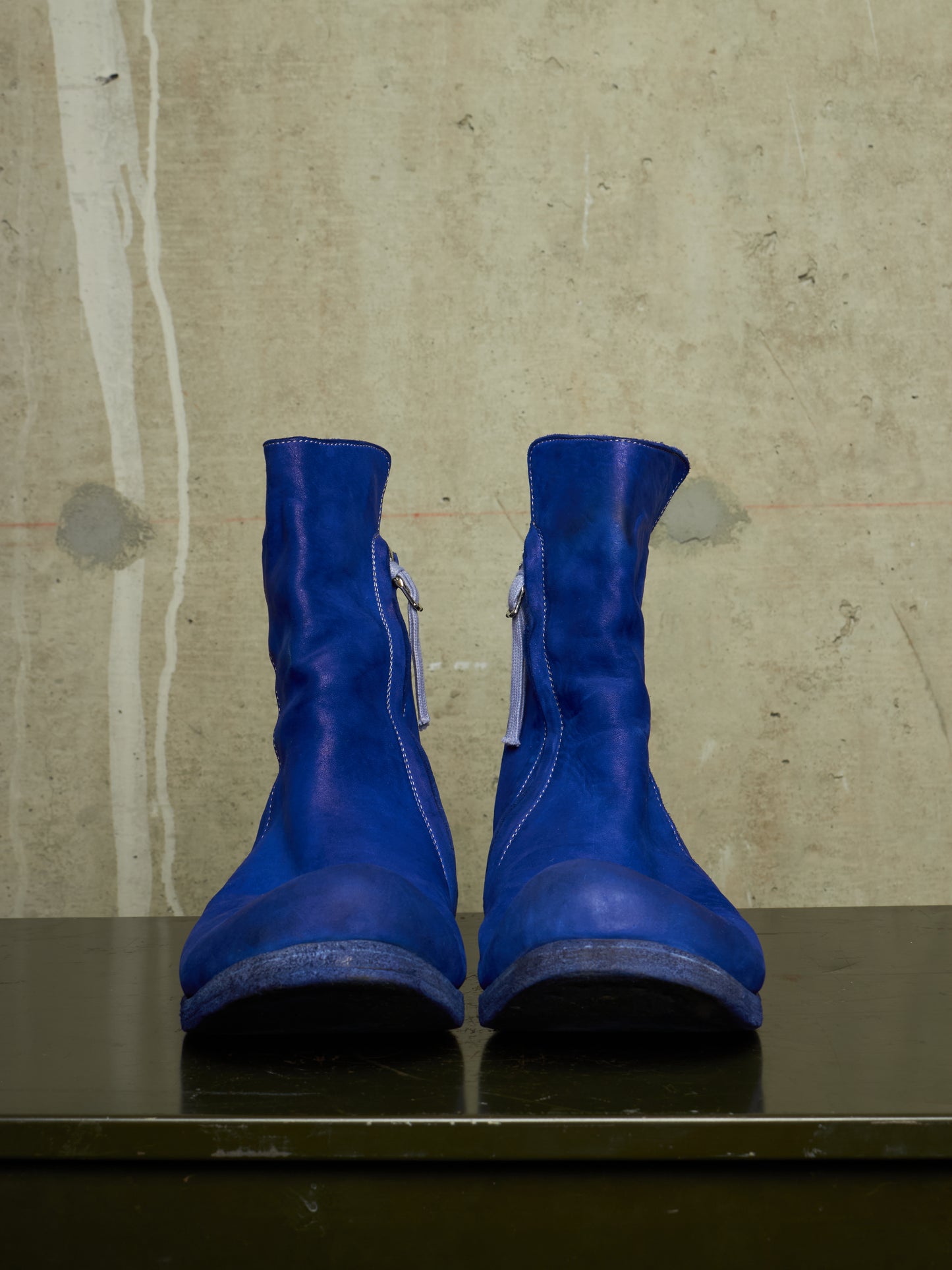 A pair of bright royal blue leather ankle boots with side zippers, displayed on a dark reflective surface against a textured concrete wall.
