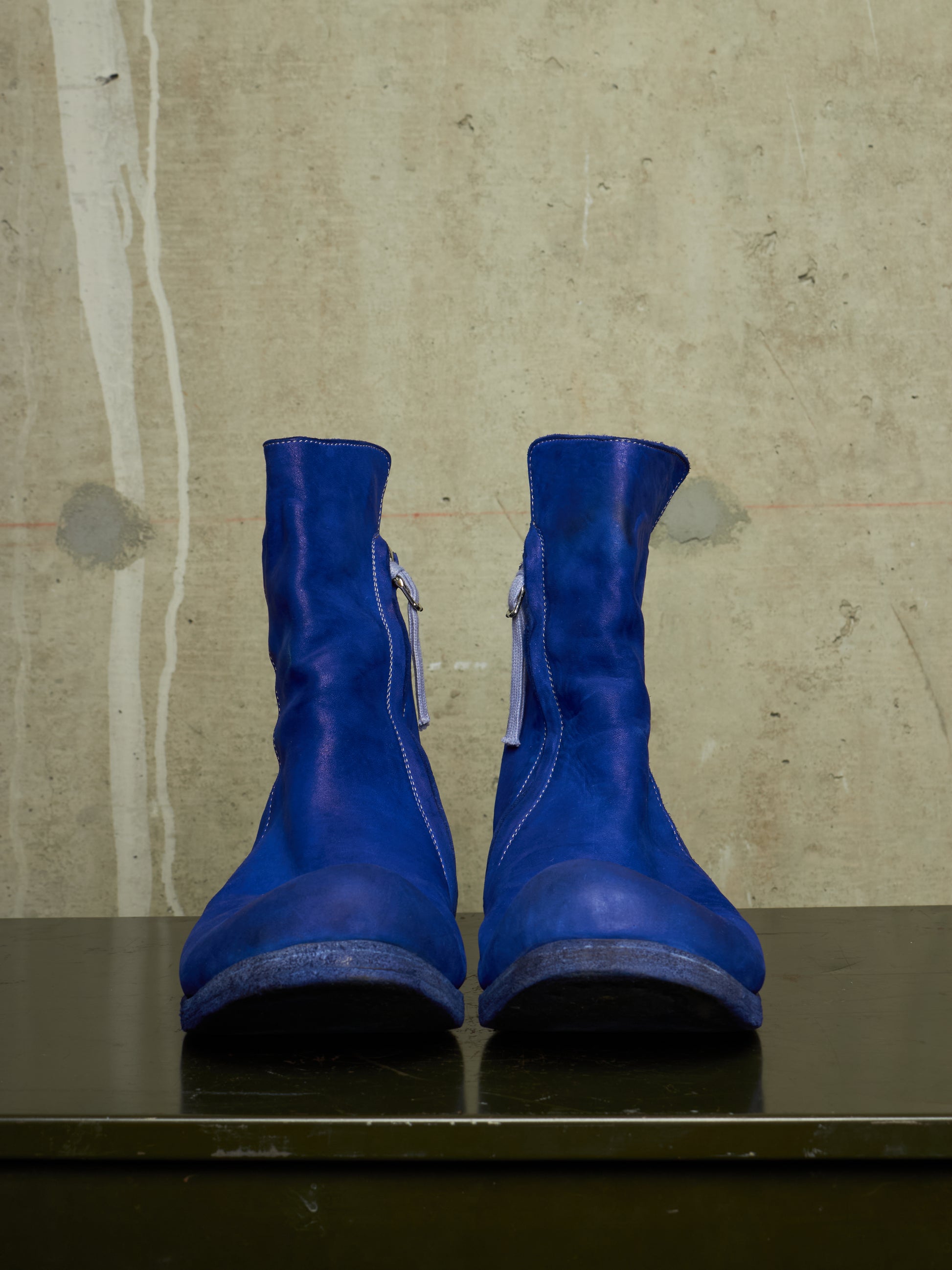 A pair of bright royal blue leather ankle boots with side zippers, displayed on a dark reflective surface against a textured concrete wall.