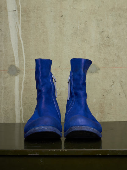 A pair of bright royal blue leather ankle boots with side zippers, displayed on a dark reflective surface against a textured concrete wall.