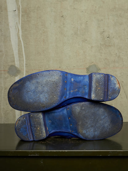 The soles of a pair of blue shoes stacked on top of each other, resting on a dark surface against a textured concrete wall.