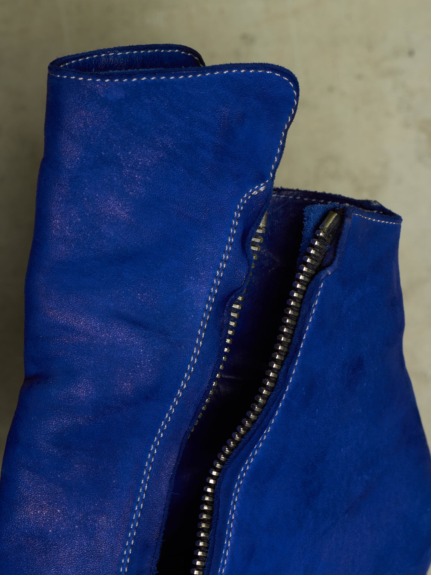 Close-up of a blue suede boot with a silver zipper and white contrast stitching, set against a neutral concrete background.