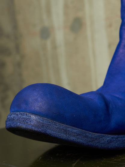 A close-up shot of a vibrant blue suede boot, showing the rounded toe and textured sole against a blurred beige background.