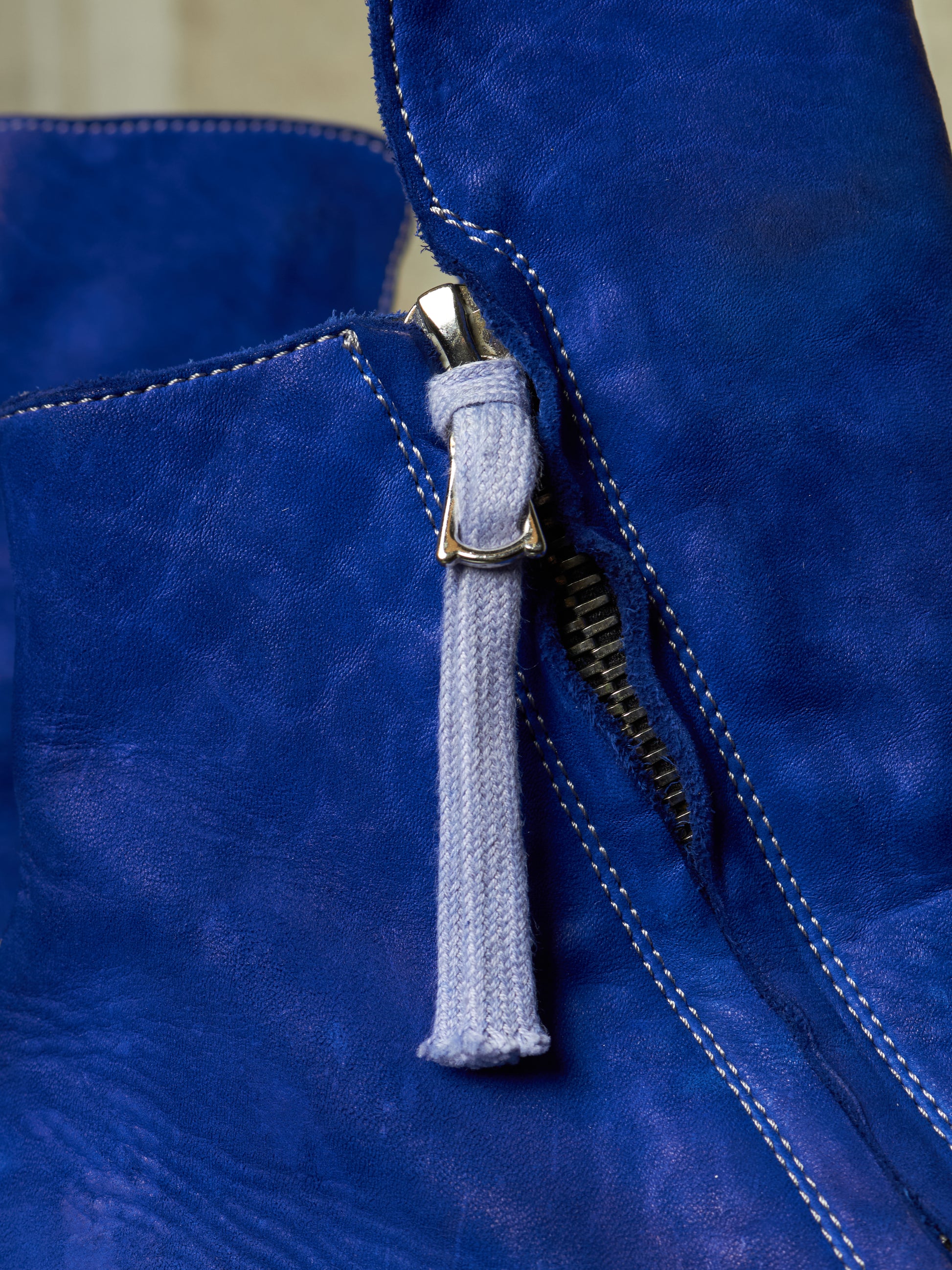 Close-up of a blue suede or leather zipper detail with a light gray fabric pull tab and gold-tone metal hardware.