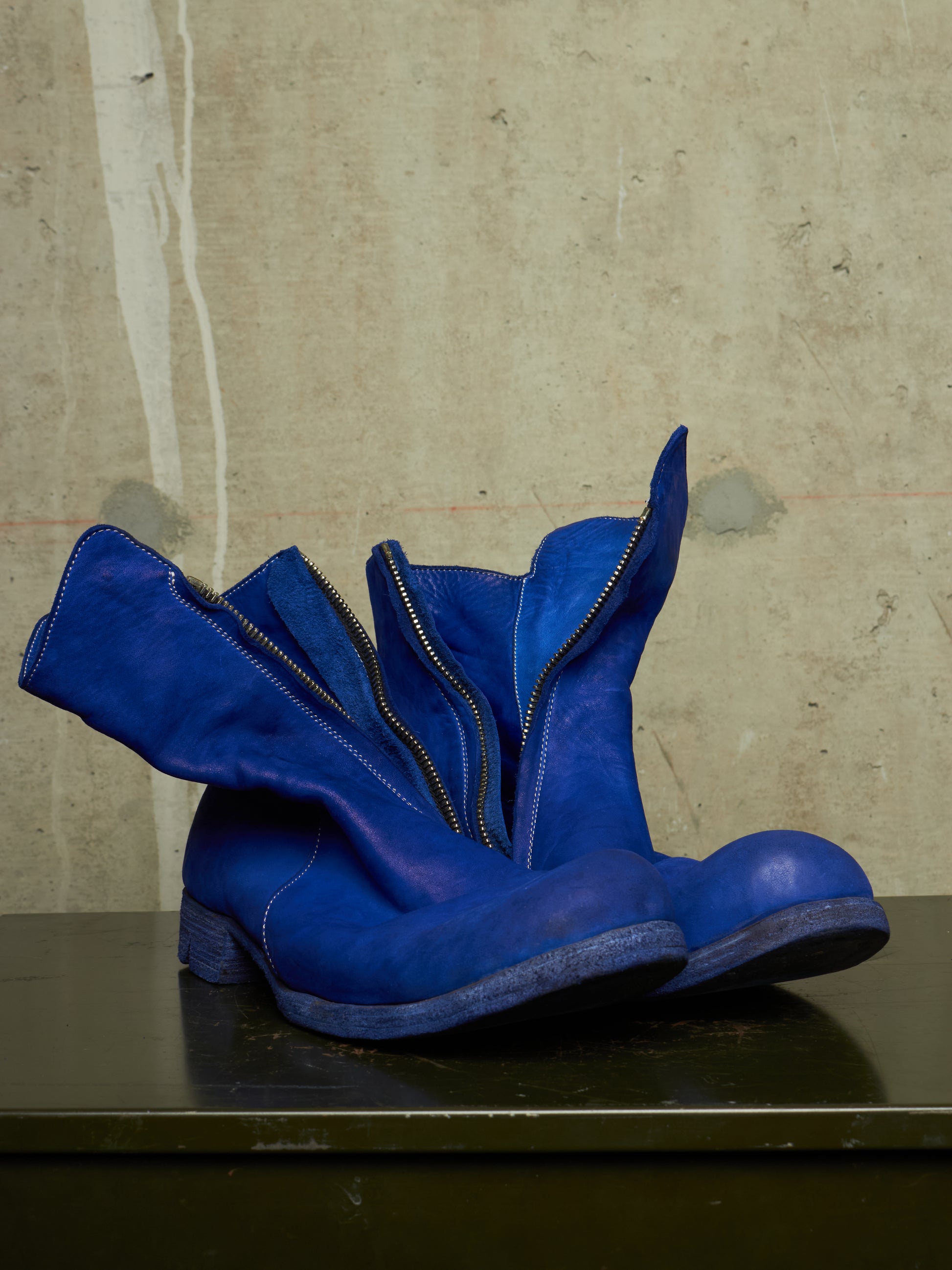 A pair of bright blue suede ankle boots with a unique flared zipper design, resting on a dark surface against a textured concrete wall.