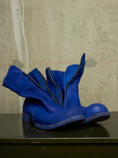A pair of bright blue suede ankle boots with a unique flared zipper design, resting on a dark surface against a textured concrete wall.