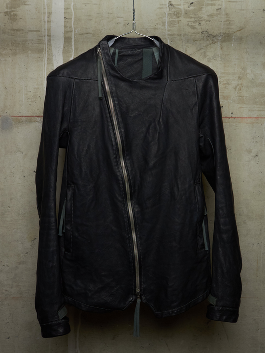 A black leather jacket with an asymmetrical zipper, hanging on a wire hanger against a textured concrete wall.