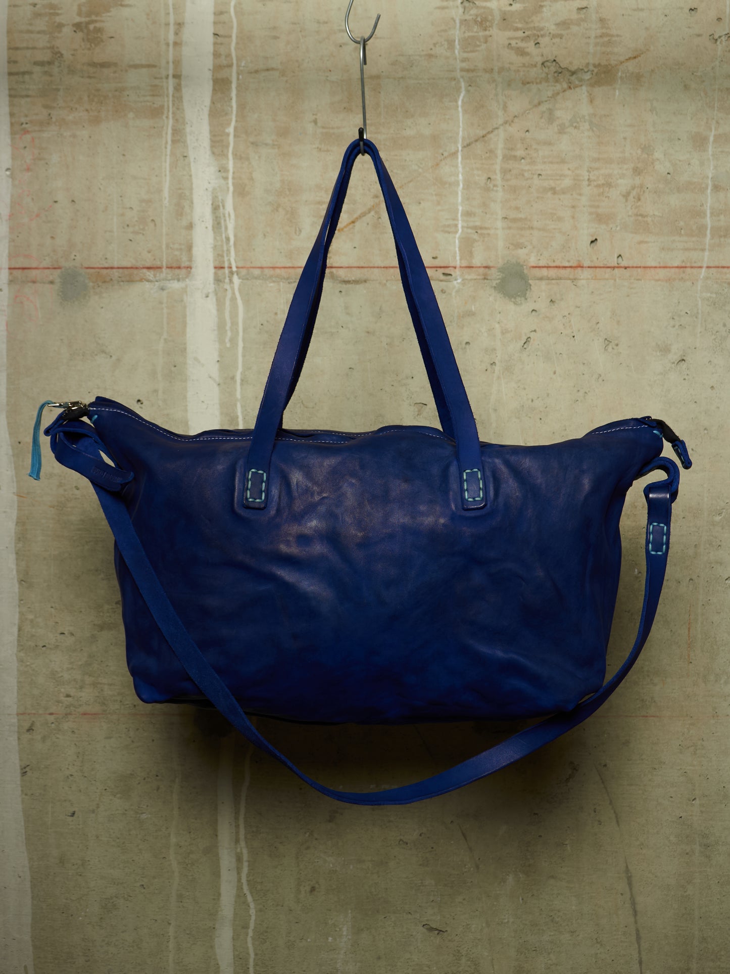 A dark blue leather duffel bag with matching shoulder straps, hanging from a metal hook against a textured concrete wall.