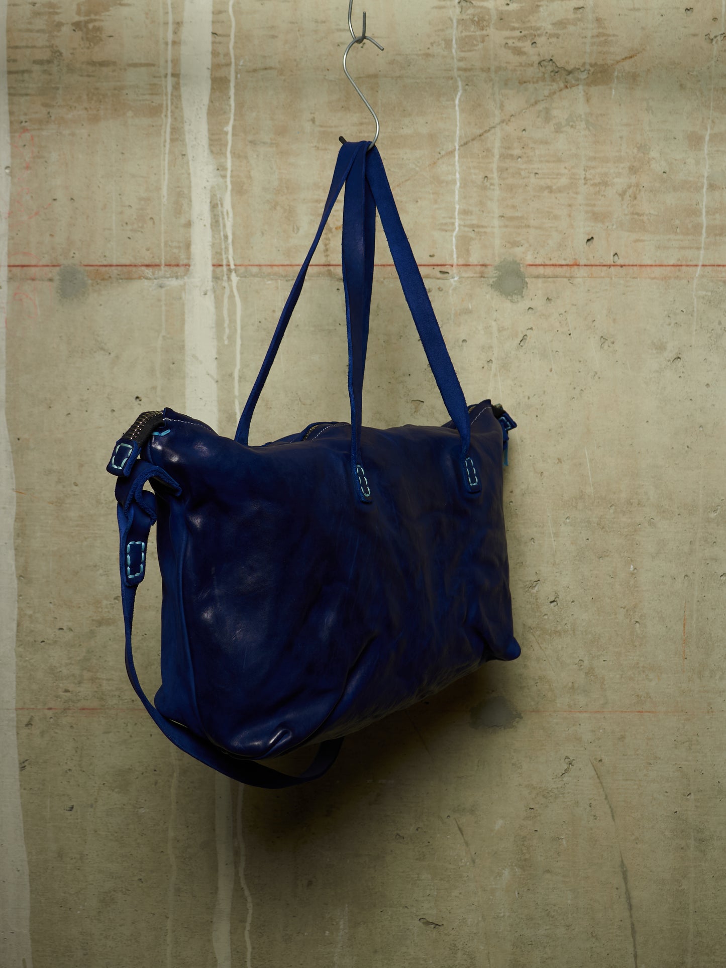 A dark blue leather duffel bag hanging from a metal hook against a textured concrete wall.