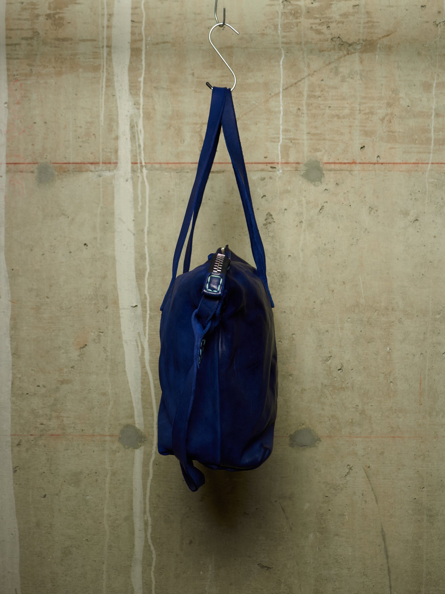 A dark blue nylon duffel bag hanging from a metal hook against a textured concrete wall.