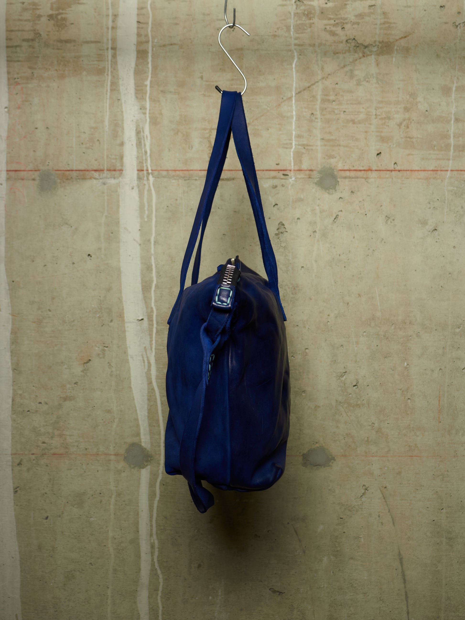 A dark blue nylon duffel bag hanging from a metal hook against a textured concrete wall.