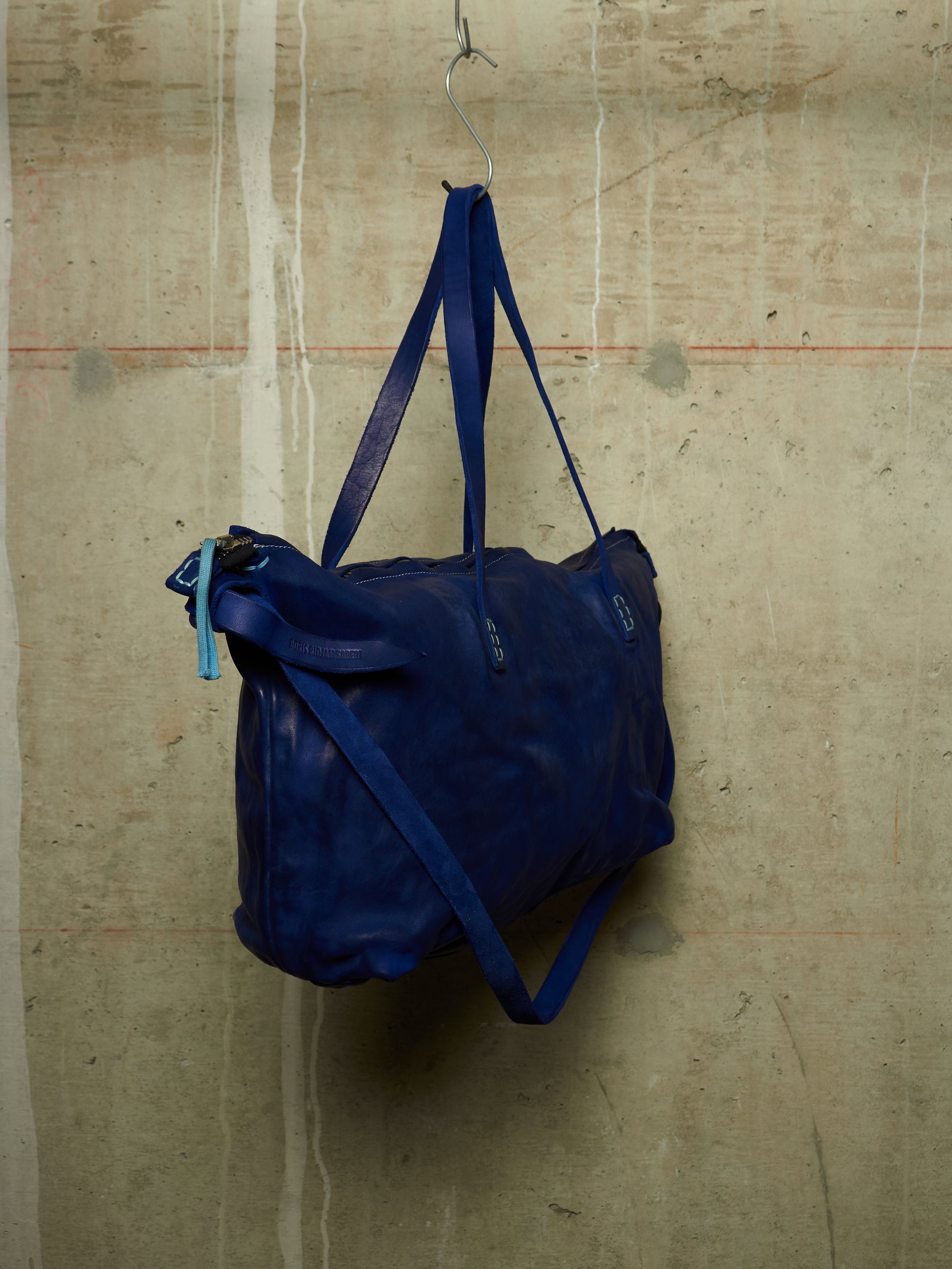 A dark blue leather duffel bag hanging from a metal hook against a textured concrete wall.