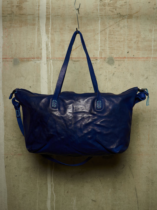 A large, crinkled dark blue leather duffel bag hanging from a metal hook against a textured concrete wall.