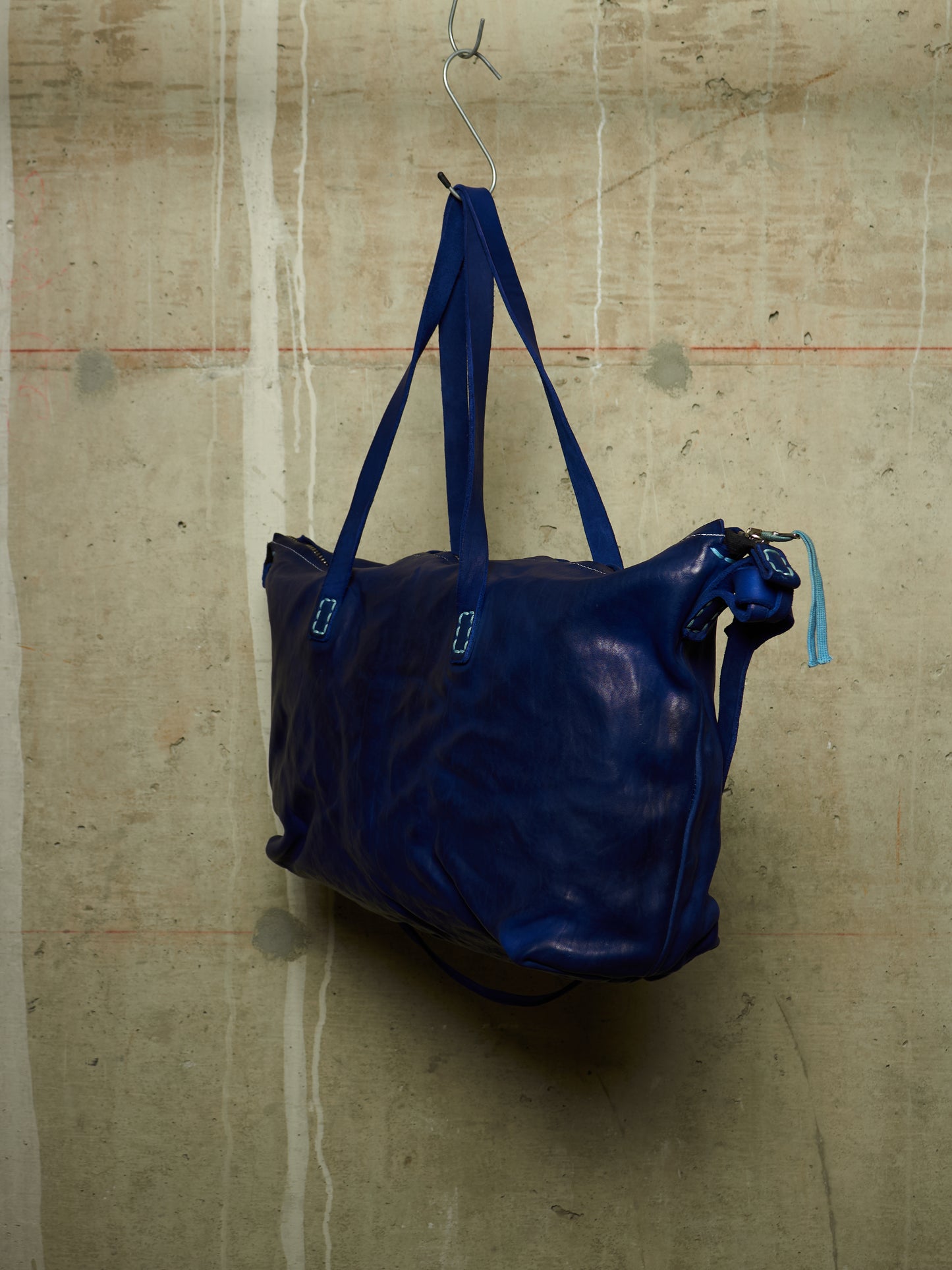 A dark blue leather tote bag with matching blue shoulder straps, hanging from a metal hook against a textured concrete wall.