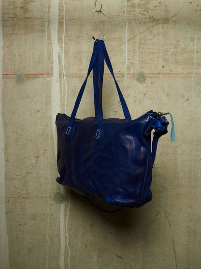 A dark blue leather tote bag with matching blue shoulder straps, hanging from a metal hook against a textured concrete wall.