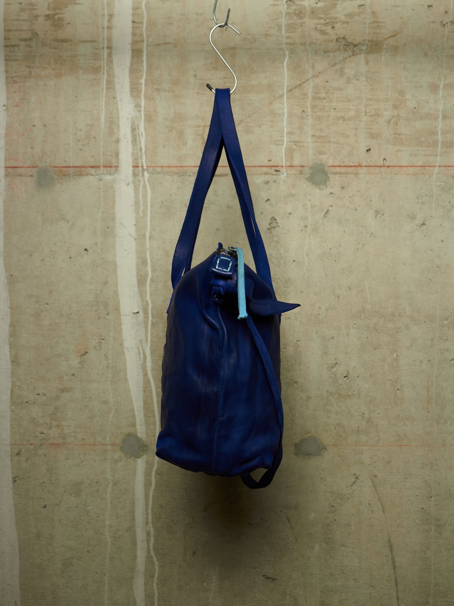 A dark blue leather duffel bag hanging from a metal hook against a textured concrete wall.