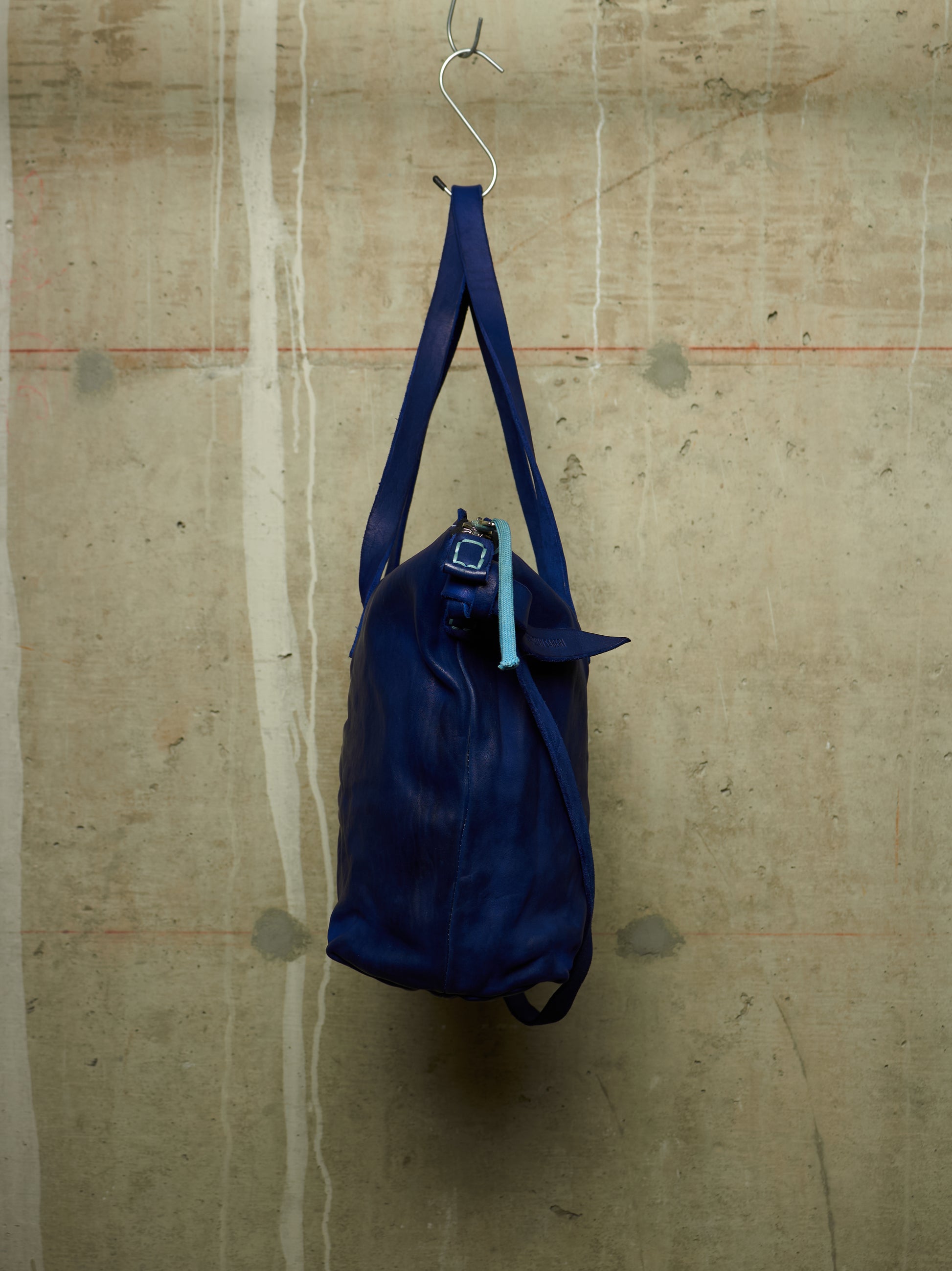 A dark blue leather duffel bag hanging from a metal hook against a textured concrete wall.