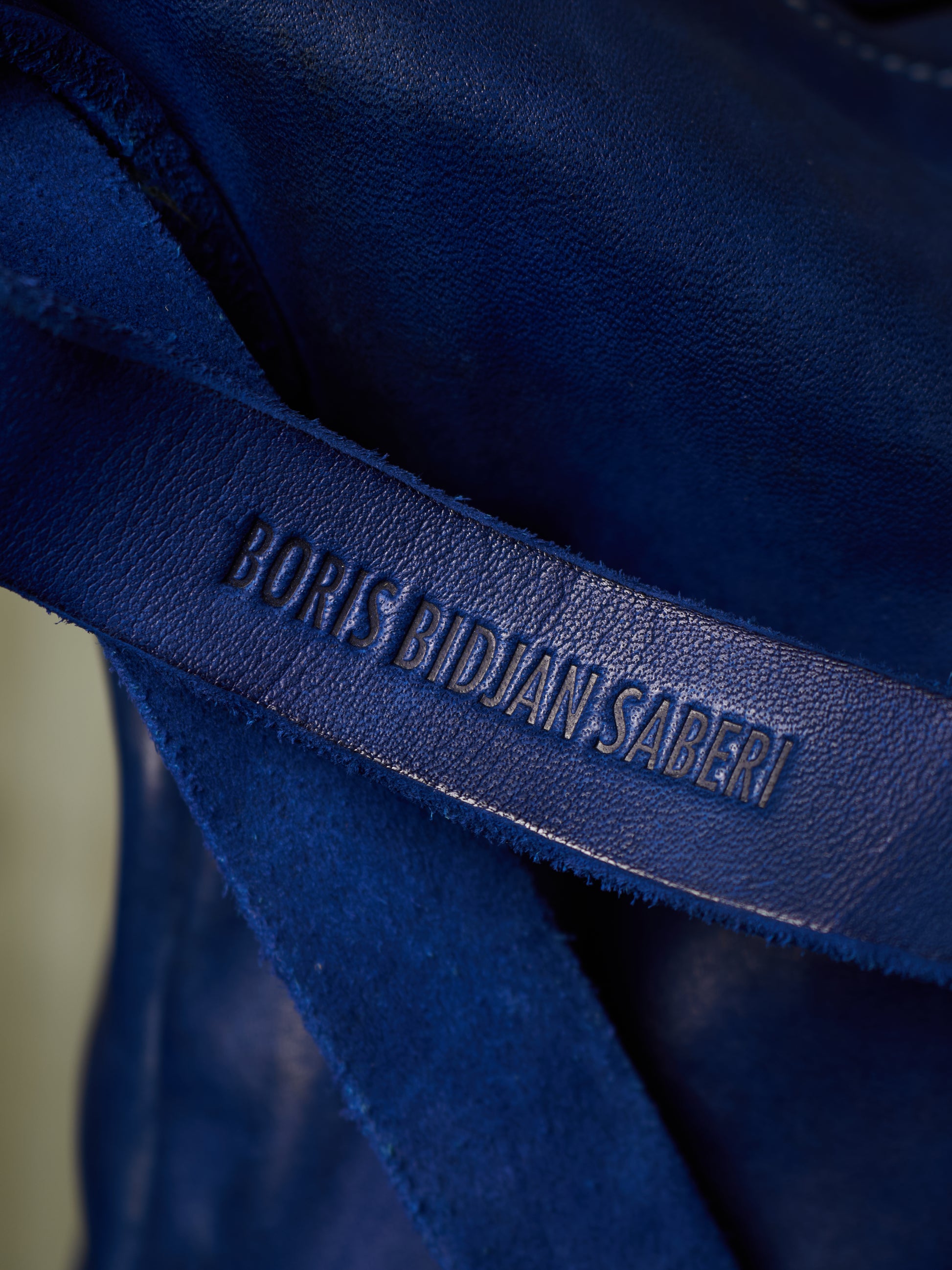 Close-up of a dark blue leather strap with the debossed brand name 'BORIS BIDJAN SABERI' on a textured blue fabric background.