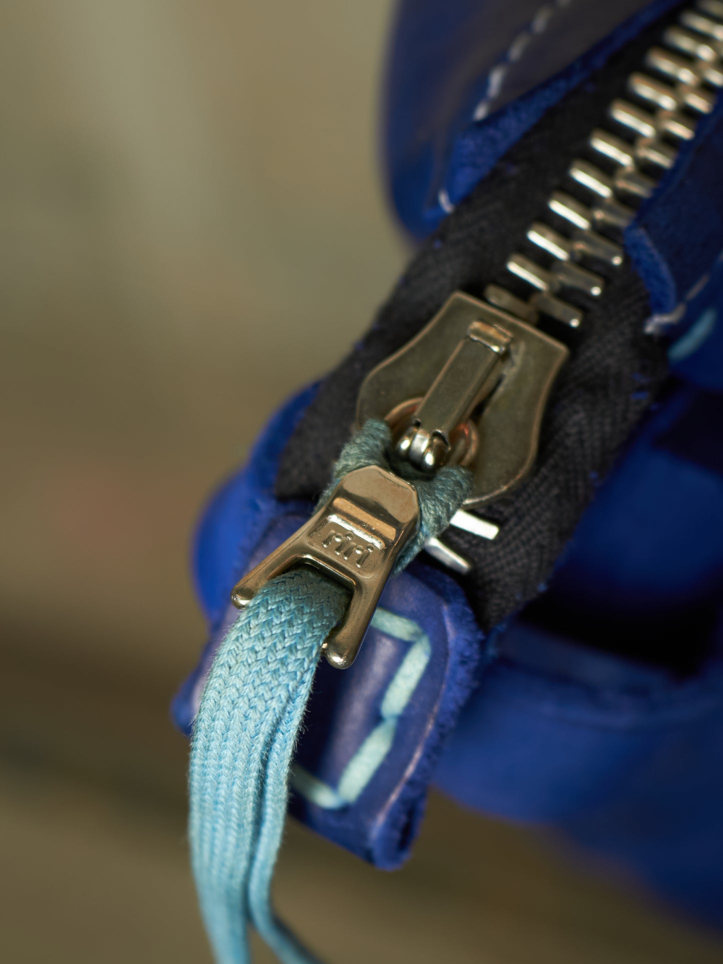 Close-up of a blue fabric zipper with a silver metal pull and a light blue woven loop tab attached to the zipper pull.