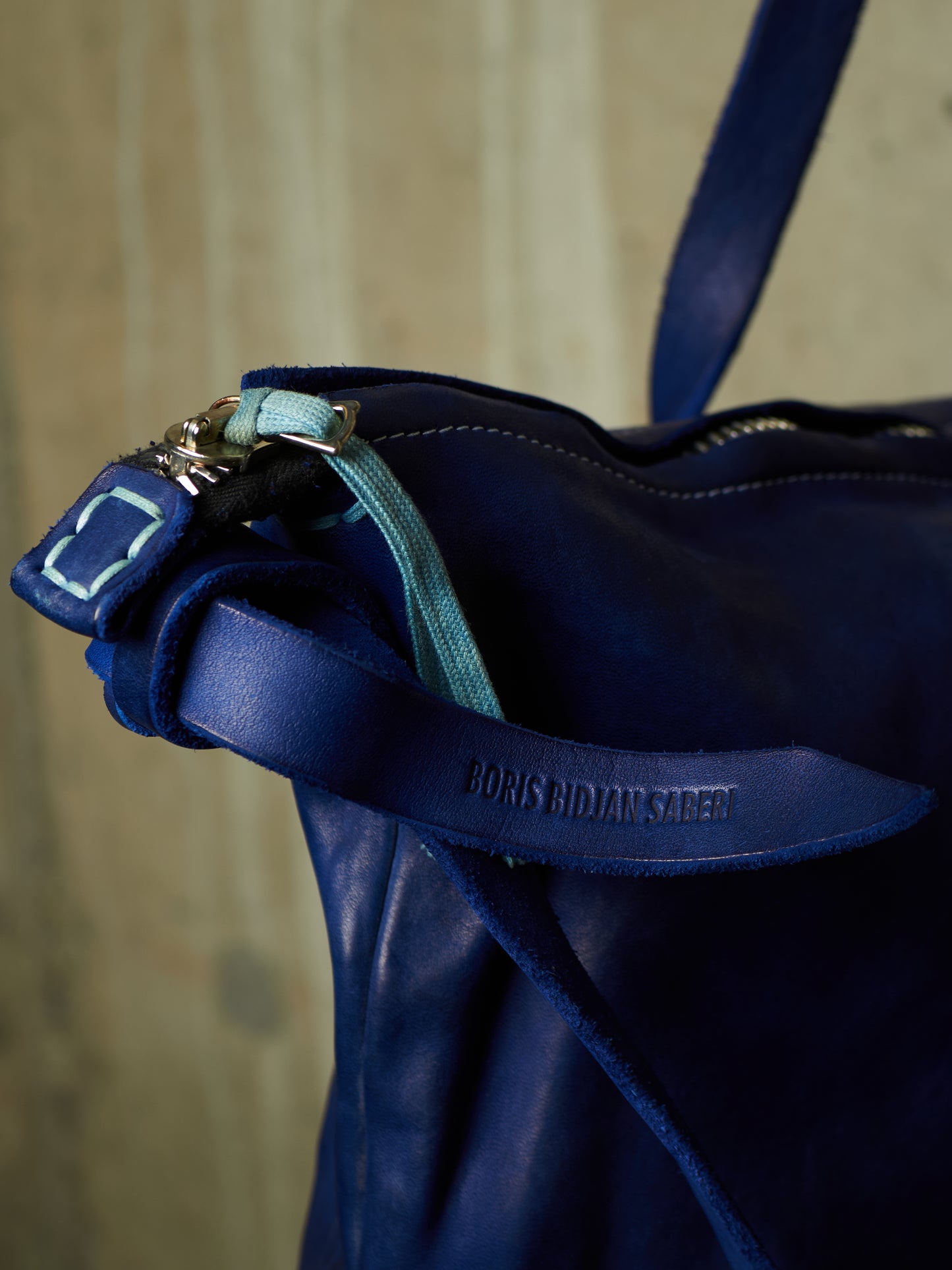 Close-up of a dark blue leather bag with a matching leather strap embossed with the brand name BORIS BIDAR SADER, featuring a light blue zipper pull and silver hardware.
