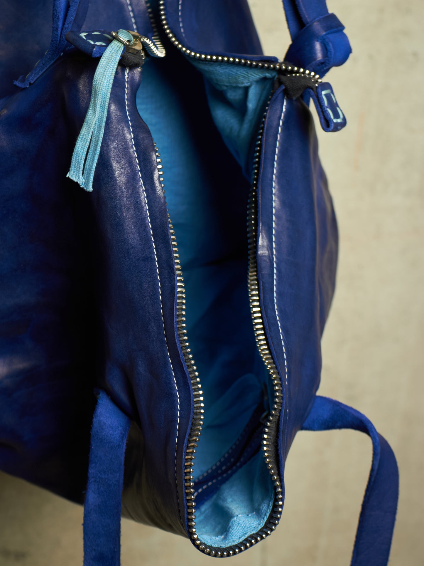 Close-up of a blue leather bag with an open zipper revealing a lighter blue interior, featuring silver-tone metal hardware and a light blue tassel zipper pull.