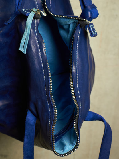 Close-up of a blue leather bag with an open zipper revealing a lighter blue interior, featuring silver-tone metal hardware and a light blue tassel zipper pull.