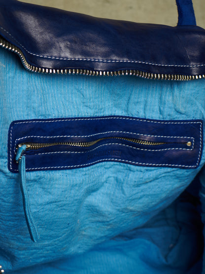 Close-up of a blue leather and crinkled fabric bag with a dark blue flap, gold-toned zipper, and white contrast stitching.