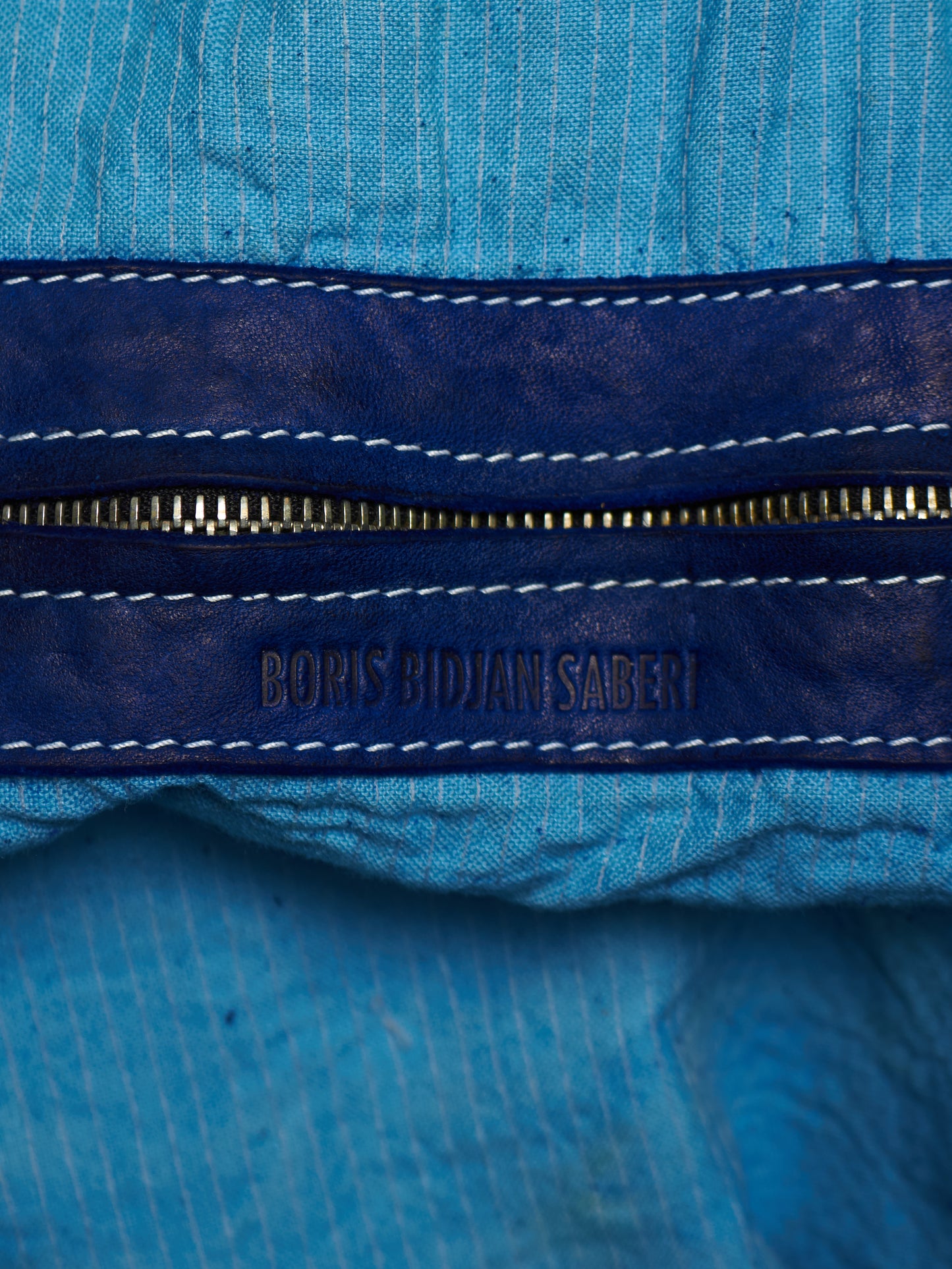 Close-up of a blue ribbed fabric garment with a dark navy leather zipper trim and white contrast stitching. The zipper pull tab is embossed with the brand name 'BORIS BIDJAN SABERI'.