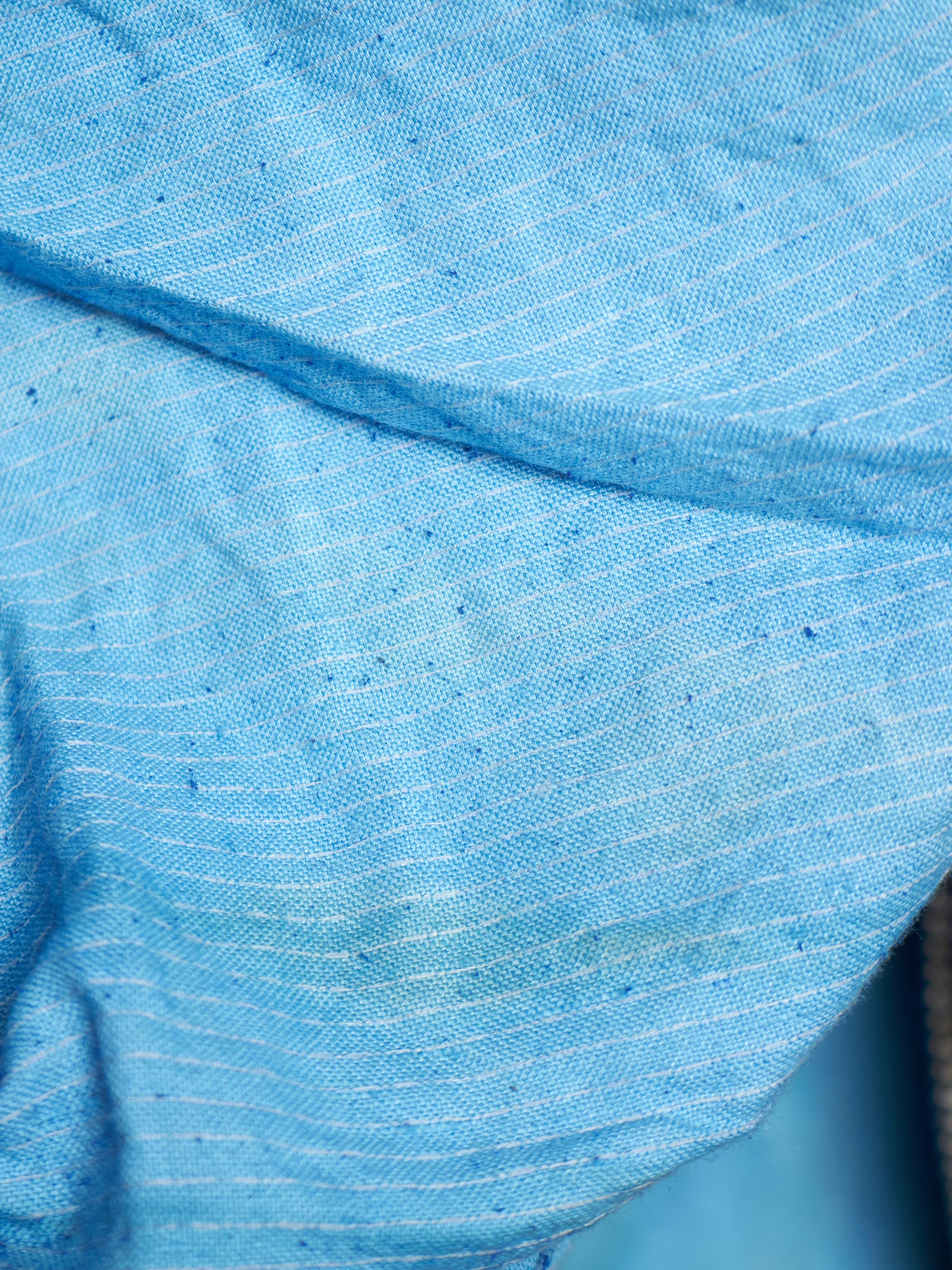 A close-up of light blue fabric with a fine white stripe pattern and a slightly textured weave.