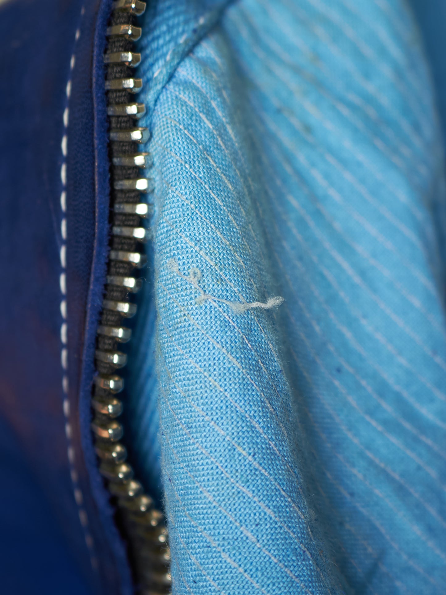 Close-up of a blue zippered garment showing the silver metal zipper, dark blue outer fabric with white stitching, and a lighter blue striped inner lining.