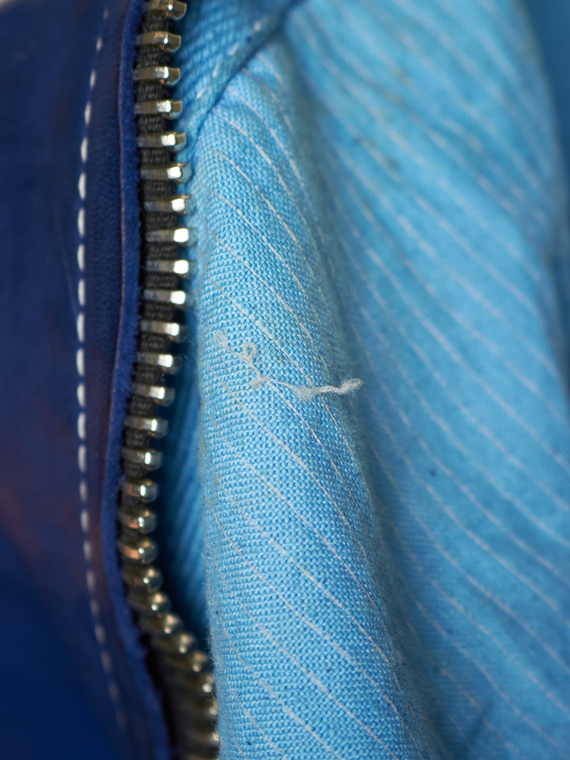Close-up of a blue zippered garment showing the silver metal zipper, dark blue outer fabric with white stitching, and a lighter blue striped inner lining.