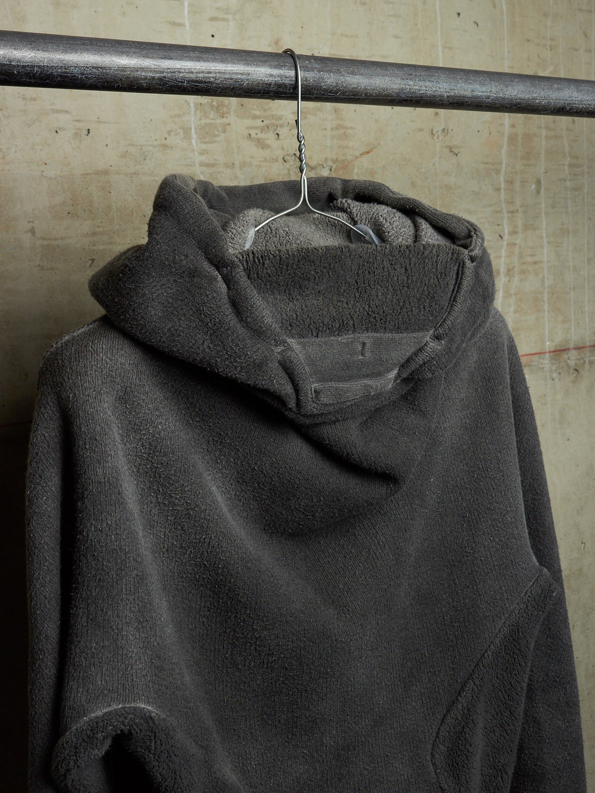 A dark gray fleece hoodie hanging on a metal hanger from a horizontal metal rod, set against a textured beige concrete wall.