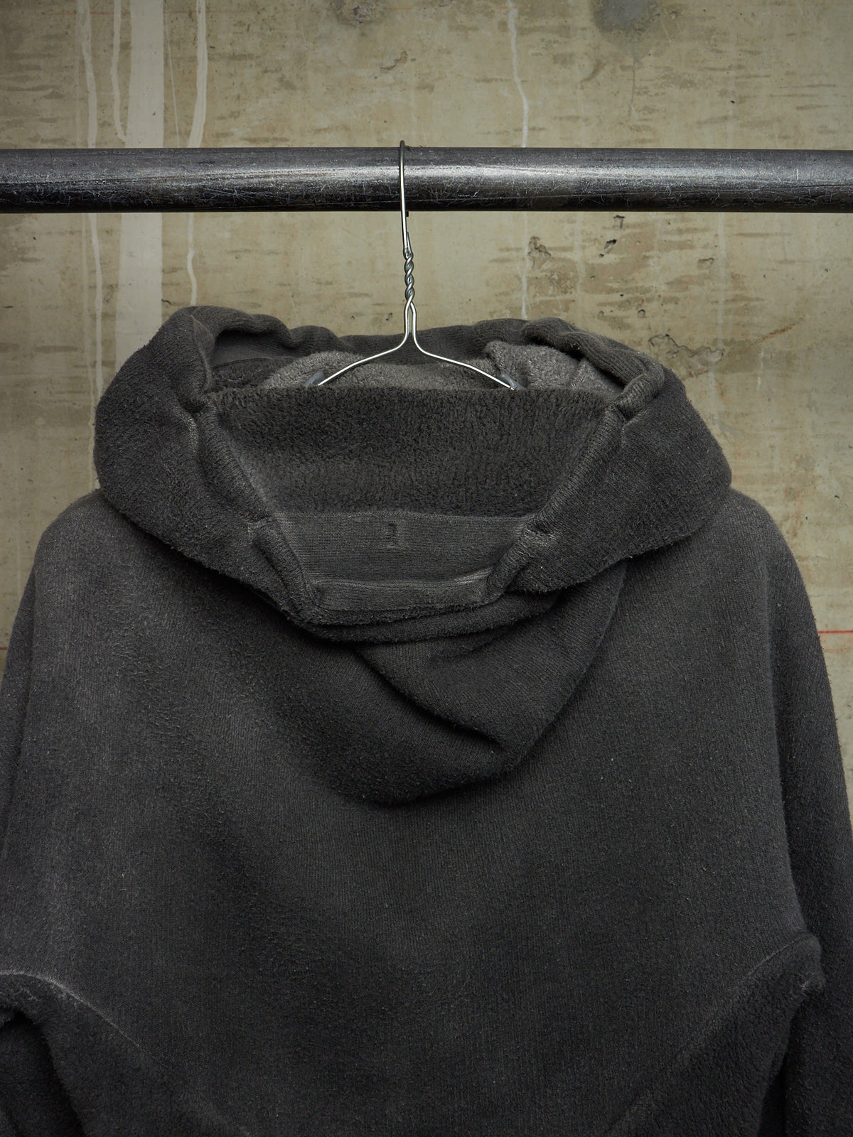 A dark charcoal gray hooded sweatshirt hanging from a metal hanger on a horizontal rod against a textured concrete wall.