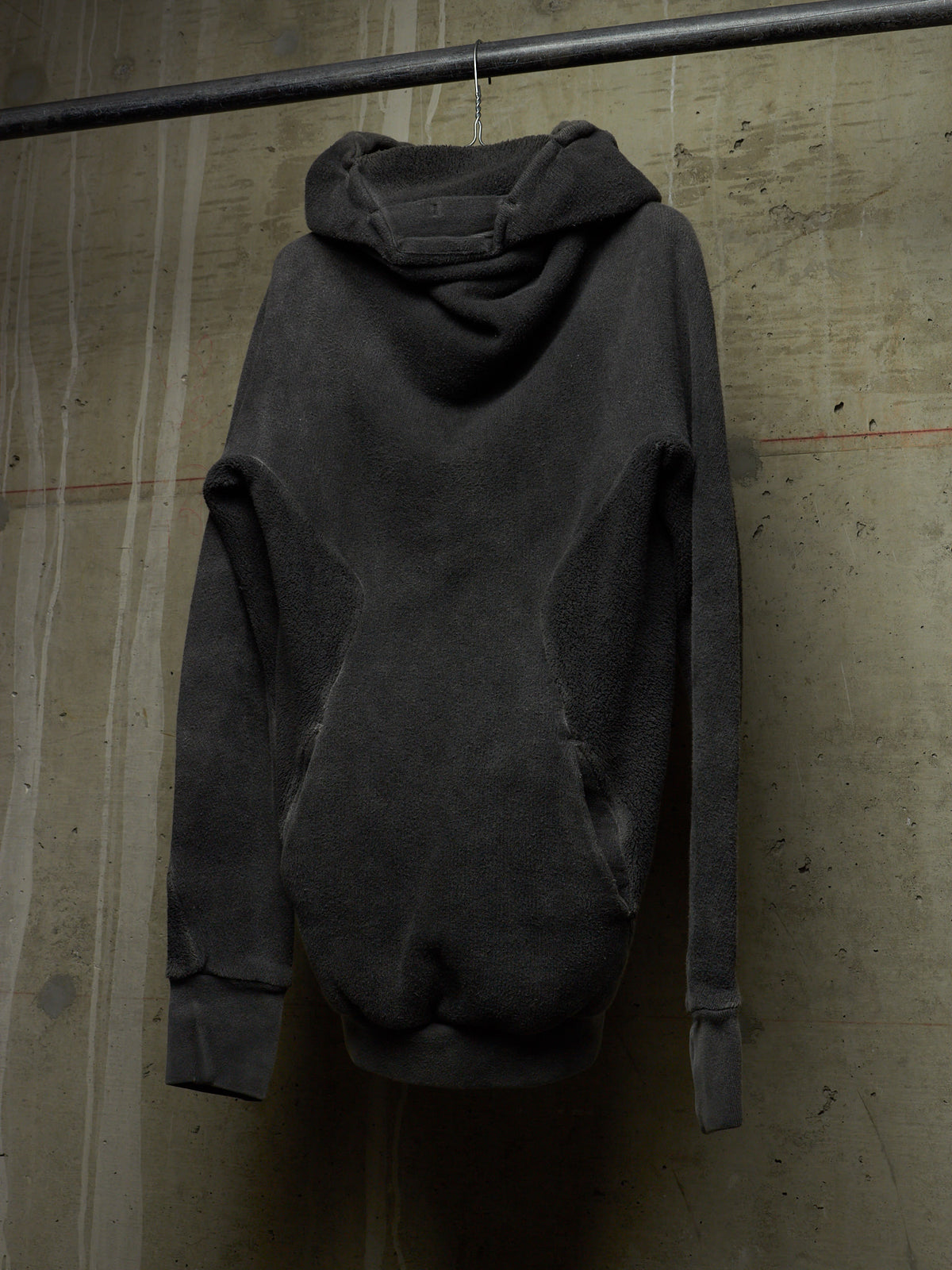 A dark charcoal gray oversized hooded sweatshirt hanging from a metal rod against a textured concrete wall.