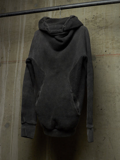 A dark charcoal gray oversized hooded sweatshirt hanging from a metal rod against a textured concrete wall.