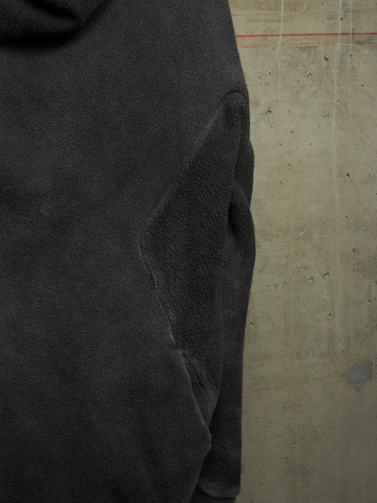 Close-up of a dark charcoal gray fleece or terry cloth garment, showing the fabric texture and a seam, set against a concrete wall background.